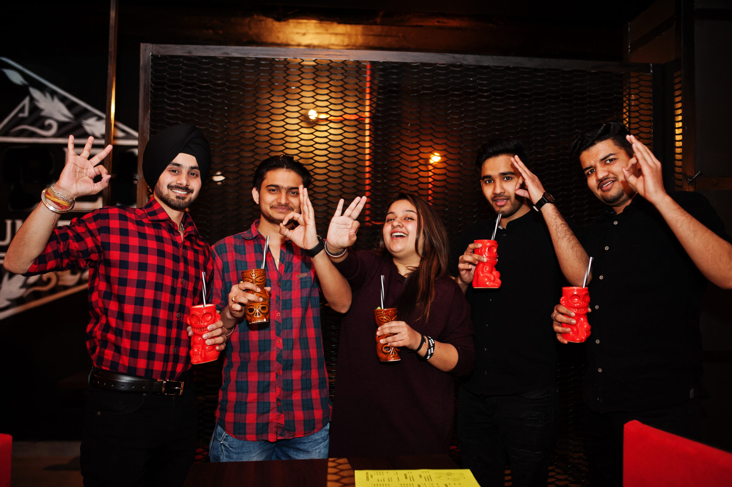 Group of indian friends having fun and rest at night club, drinking cocktails and show ok fingers together.