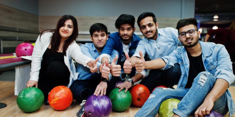 Group of five south asian peoples having rest and fun at bowling club. Holding bowling balls, sitting on alley and show thumb up hands.