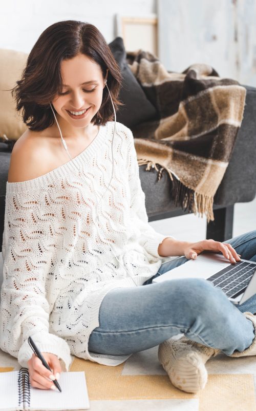 cheerful girl with earphones studying online with laptop and notebook at home